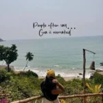 Goa Captions Instagram: Vertical photograph of a person wearing a yellow cap, a dark top, and a white skirt standing at a wooden railing, looking out toward a tropical beach and ocean view. Centered over the sky and water is black handwritten-style text that reads "People often say... Goa is overrated."