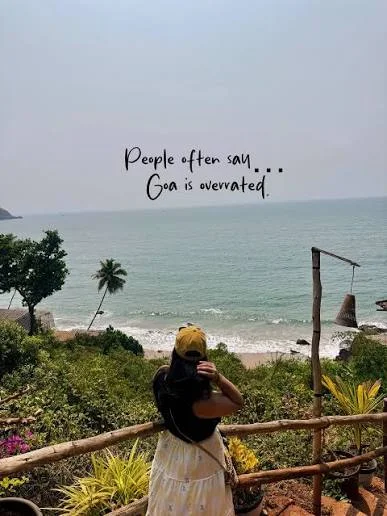Goa Captions Instagram: Vertical photograph of a person wearing a yellow cap, a dark top, and a white skirt standing at a wooden railing, looking out toward a tropical beach and ocean view. Centered over the sky and water is black handwritten-style text that reads "People often say... Goa is overrated."